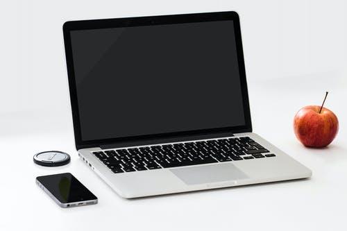 Auki oleva kannettava tietokone jonka vasemmalla puolella matkapuhelin ja oikealla puolella punainen omena.