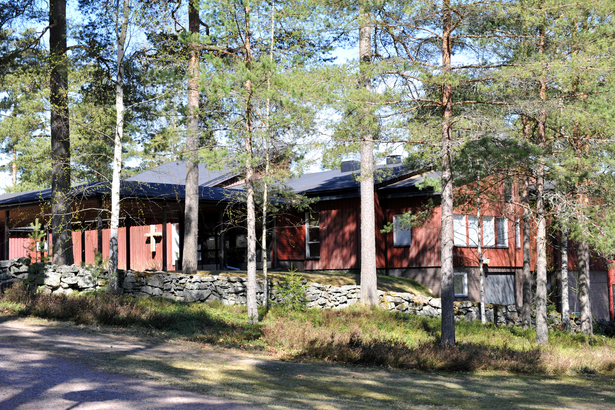 Punainen talo, mäntyjen keskellä, missä sijaitsee Dragsfjärdin seurakuntakoti ja seurakunnan kanslia. Etualalla kivimuuri.